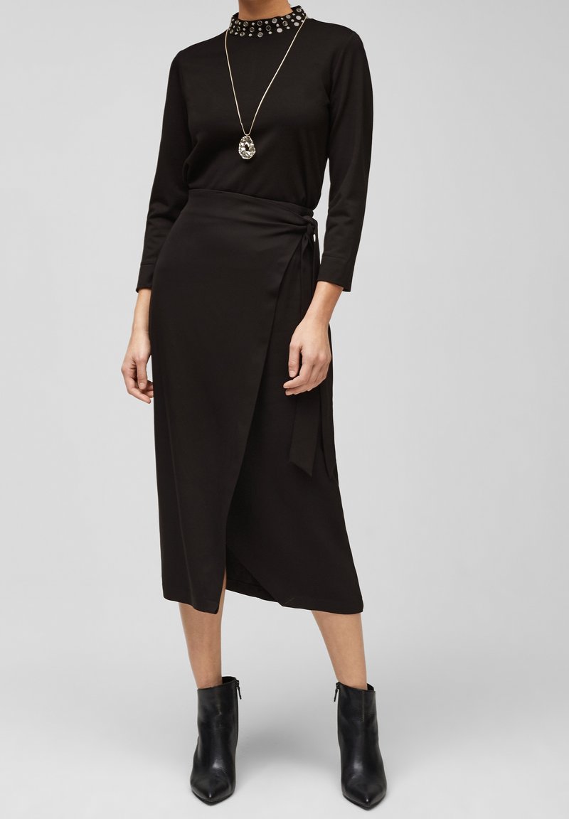 Czarny, długi rękaw, bluzka z wysokim kołnierzem ozdobiona ćwiekami, połączona z czarną spódnicą typu wrap związaną w talii. Botki na obcasie sięgające do kostek.