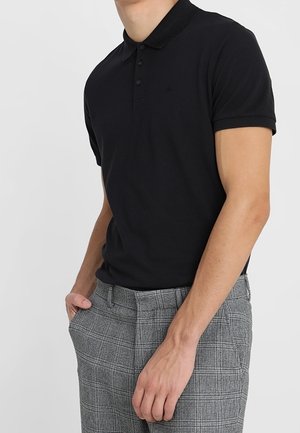 Mand iført en sort poloshirt med korte ærmer og grå ternede bukser, stående med den ene hånd i lommen mod en ensfarvet baggrund.