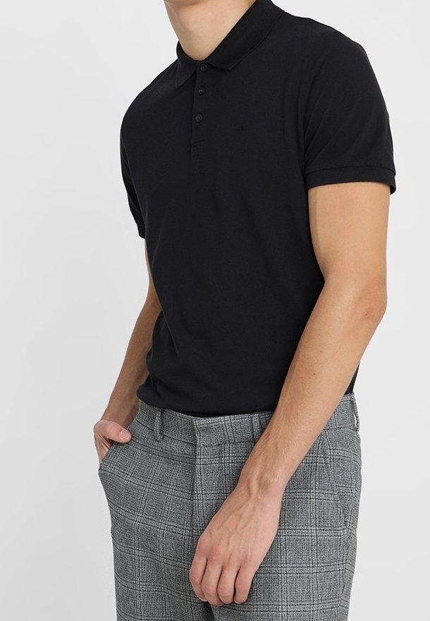 Homme portant un polo noir à manches courtes et un pantalon gris à carreaux, debout avec une main dans la poche sur un fond uni.
