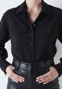 Camicia nera con bottoni, realizzata in un tessuto testurizzato, dotata di due tasche sul petto e colletto classico. Indossata con pantaloni in pelle nera a vita alta.