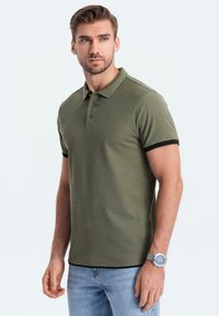 Olivově zelené polo triko s černými detaily, krátké rukávy a knoflíková lišta se třemi knoflíky. Doplňují ho světle modré džíny a stříbrné hodinky.
