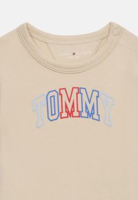 Μπεζ βαμβακερό μπλουζάκι με στρογγυλό λαιμό και κοντά μανίκια. Διαθέτει πολύχρωμο λογότυπο "TOMMY" σε κόκκινη, μπλε και ανοιχτό μπλε κέντημα.