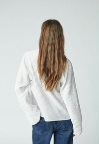 Cămașă albă, texturată, cu mâneci lungi, cu o croială relaxată și umeri lăsați, asortată cu blugi din denim albastru. Design minimalist, fără modele vizibile.