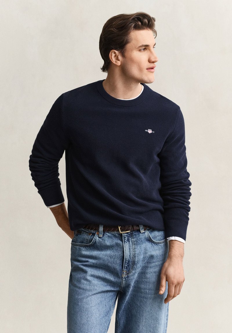 Jeune homme aux cheveux foncés portant un pull bleu marine et un jean bleu clair regarde sur le côté avec la main gauche dans la poche.