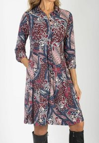 Kobieta ubrana w sukienkę w długości do kolan, w wzory z maroonowymi, niebieskimi i białymi kwiatowymi oraz geometrycznymi wzorami, w połączeniu z czarnymi kozaczkami do kolan.