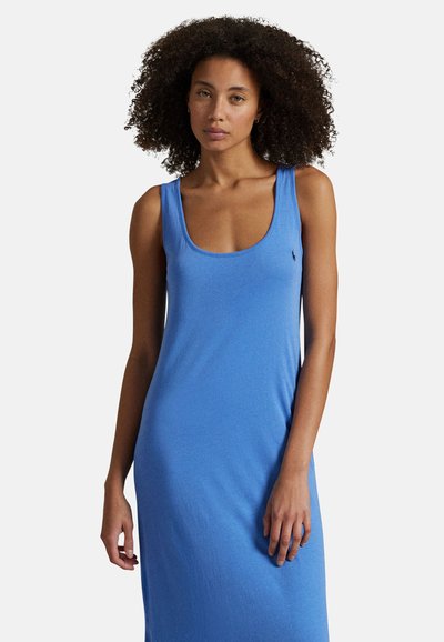 Kobieta o kręconych włosach, ubrana w niebieską sukienkę bez rękawów z zaokrąglonym dekoltem, stojąca na tle jednolitego jasnego tła.