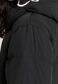 Černá péřová bunda se štepovaným vzorem, hladkým texturem a kapucí na šňůrku s bílým logem na zadní straně.