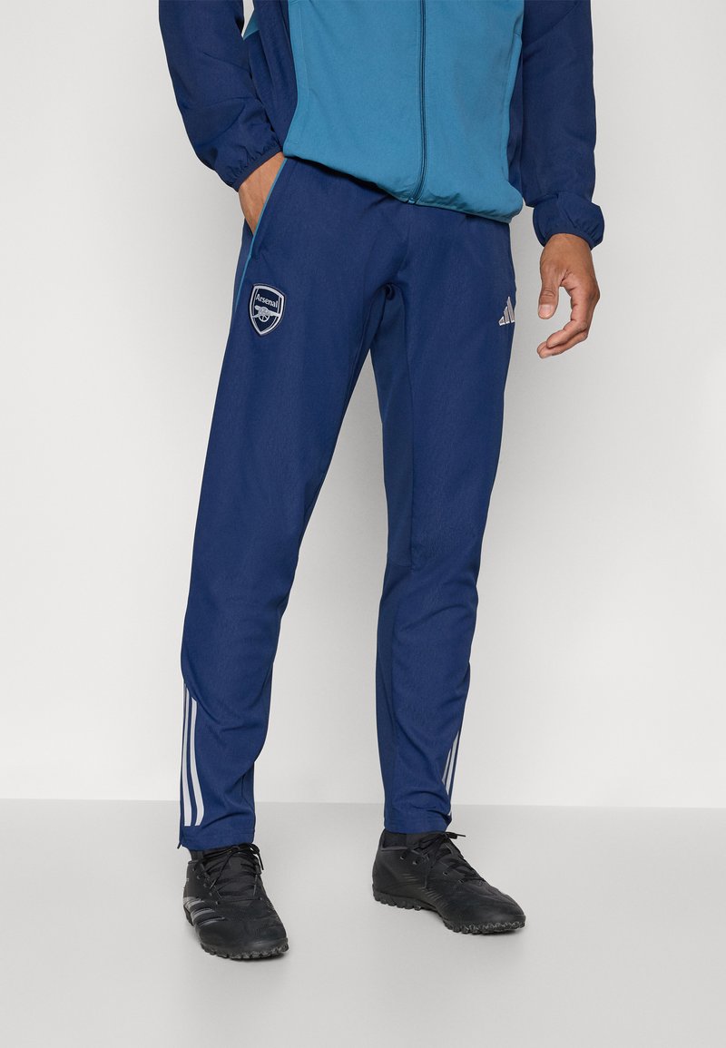Marine atletiske bukser lavet af glat stof; har hvide Adidas-striber på siderne og et Arsenal-logo på den venstre lår.
