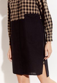 Czarna sukienka koszulowa z wierzchnim materiałem w kratkę w odcieniach beżu i czerni. Posiada rozcięcie z boku oraz kieszeń na piersi z zapięciem na zatrzask.