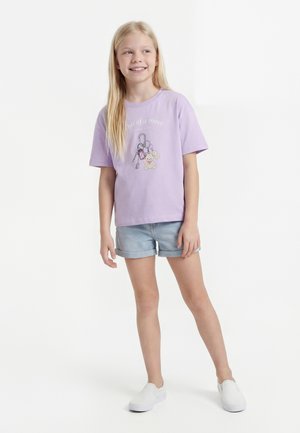 Lány hosszú szőke hajjal, aki levendulaszínű pólót visel kulcs és mackó grafikával, világos farmer rövidnadrágot és fehér bebújós cipőt, mosolyogva, jobb oldalt nézve.