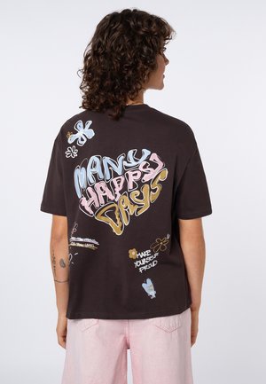 Person with curly hair wearing a black t-shirt that reads "Many Happy Days" with colorful floral and text designs, paired with light pink pants.