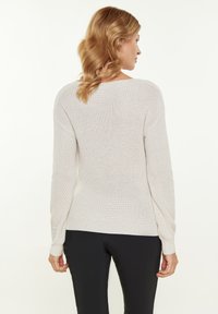 Ljus beige, stickad tröja med lång ärm, ribbat avslut och strukturerat mönster. Bärs med svarta, figurnära byxor, sedd bakifrån.