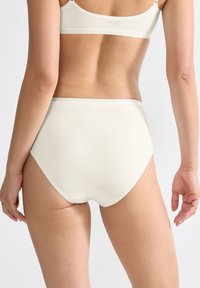 Vrouw van achteren in hoge crèmekleurige onderbroek en bijpassende bh op een effen lichte achtergrond.