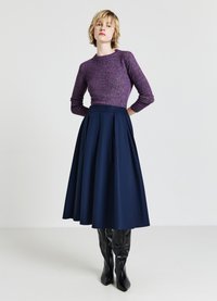 Lila Wollmischpullover mit langen Ärmeln, kombiniert mit einem marineblauen, plissierten Midirock und schwarzen, oberschenkelhohen, spitzen Stiefeln. Flacher Hintergrund.