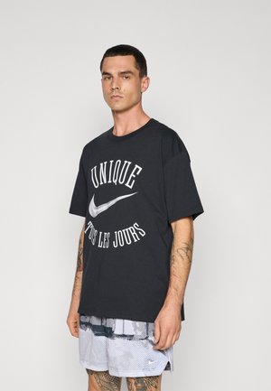 Mann mit kurzen dunklen Haaren, der ein schwarzes, übergroßes T-Shirt mit der Aufschrift "Unique Tous Les Jours" und weißem Nike-Logo trägt, kombiniert mit hellgrauen, gemusterten Shorts.