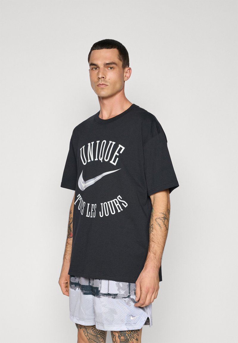 Sort t-shirt i sort bomuld med hvid tekst "UNIQUE TOUS LES JOURS" og et sølvfarvet Nike swoosh, parret med lysegrå mesh shorts.