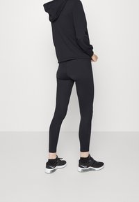 Persona vestida con sudadera negra, leggings negros y zapatillas Nike negras, de pie contra un fondo blanco liso, mirando hacia atrás.