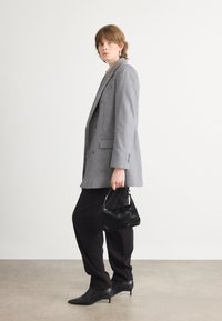 Grijze dubbelgeknoopte blazer, zwarte op maat gemaakte broek, puntige zwarte hakken en een kleine zwarte handtas met een zachte textuur. Eenvoudige achtergrond.