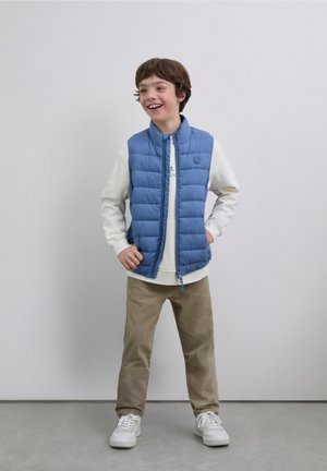 Chaleco acolchado azul claro con acolchado horizontal, cierre de cremallera y pequeño logotipo. Lleva una sudadera blanca debajo, combinado con pantalones beige y zapatillas blancas.