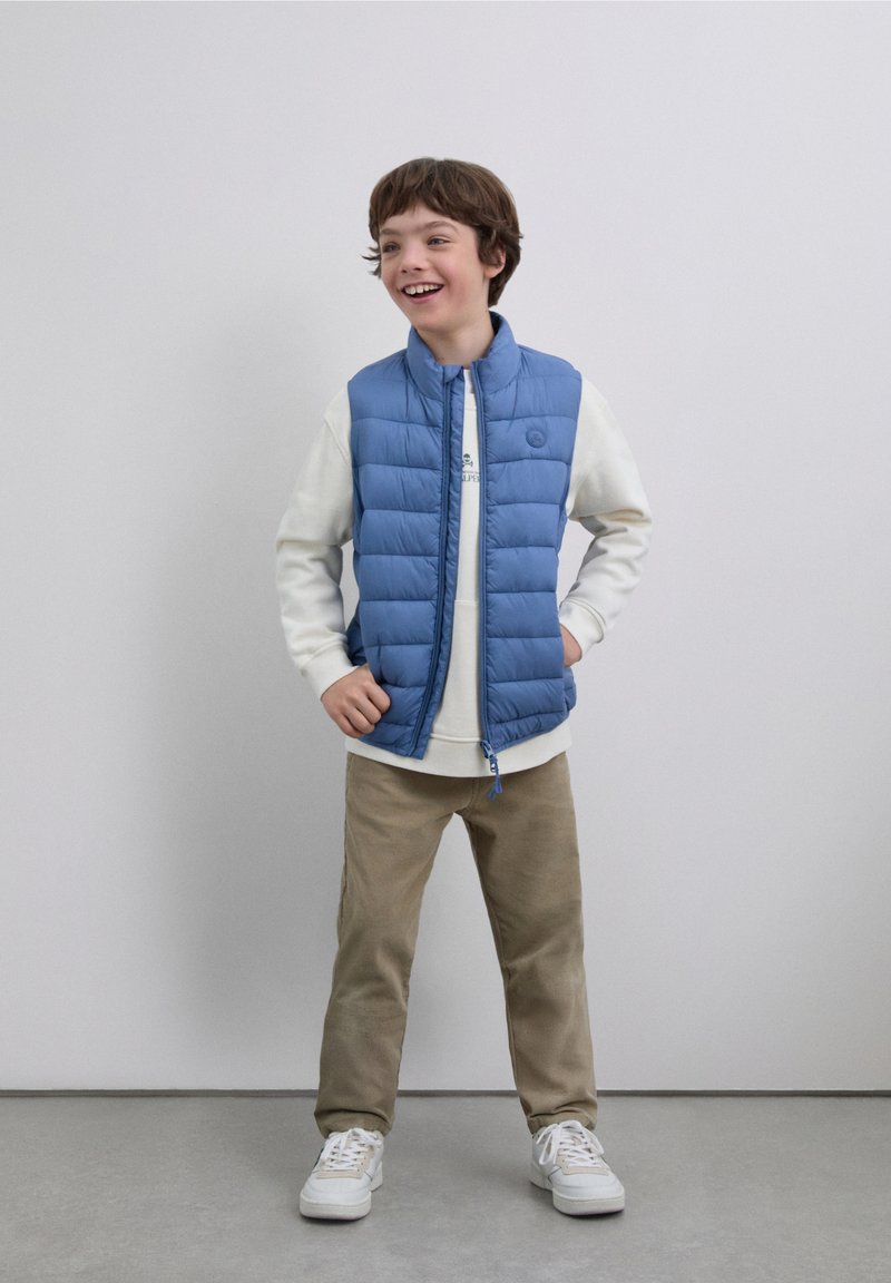 Chaleco acolchado azul claro con acolchado horizontal, cierre de cremallera y pequeño logotipo. Lleva una sudadera blanca debajo, combinado con pantalones beige y zapatillas blancas.