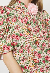 Blouse florale en tissu léger, ornée de motifs roses, rouges et verts, à manches bouffantes, avec un grand accent de fleur rose au niveau du col.