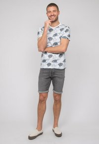 T-shirt blanc avec impression de feuilles bleu marine, associé à un short en denim gris à revers. Le modèle porte des chaussures à enfiler de couleur claire, se tenant devant un fond neutre.