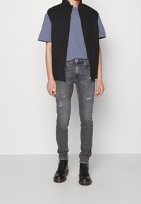 Chaleco sin mangas negro sobre una camiseta azul, combinado con jeans grises desgastados y botas negras con cordones. El conjunto presenta un ajuste moderno.