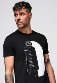 Mies, jolla on kiharaiset hiukset ja tatuointeja, pukeutuu mustaan t-paitaan, jossa on valkoinen graafinen kuva ja teksti "OMBRE menswear", katsoo sivulle tasaisen taustan edessä.