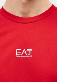 Κοντινό πλάνο ενός ατόμου που φοράει κόκκινο πουκάμισο με λευκό λογότυπο "EA7 Emporio Armani" στο στήθος.