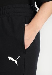 Zwarte joggingbroek van zacht materiaal, met een elastische tailleband en een wit Puma-logo op de voorkant. Zak zichtbaar aan de zijkant.