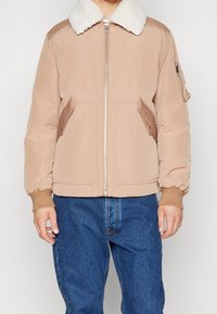 Beige bomberjacka med vit faux shearling-krage, dragkedja fram, bruna läderdetaljer och elastiska ärmslut; matchat med blå denimjeans.