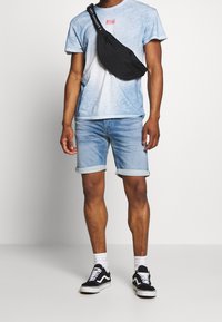 Hombre con camiseta de tie-dye azul claro, pantalones cortos de mezclilla claros enrollados, bolso cruzado negro, calcetines blancos y zapatillas negras con cordones blancos.