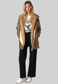 Veste matelassée dorée et brillante avec un col en fausse fourrure, portée sur un t-shirt imprimé. Un pantalon noir à large jambe et des baskets blanches complètent la tenue.