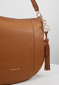 Sac à main Michael Kors en cuir cognac avec un pendentif logo doré et un pompon attaché à la bandoulière, sur un fond uni.