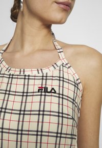Beżowy top na ramionkach w czarno-czerwony wzór w kratę. Zawiera czarną wyszywaną naszywkę z logo "Fila" w pobliżu dekoltu. Gładka struktura materiału.