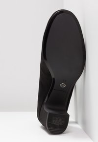 Chaussure à talon haut en daim noir avec une semelle lisse et brillante. Présente un bout arrondi et un accent argenté sur la semelle. Taille marquée 37.