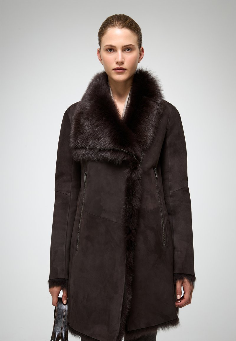 Palton maro din suede, cu un guler lat din blană, mâneci lungi și buzunare cu fermoar, având un design elegant, adaptat pe corp și o textură moale.