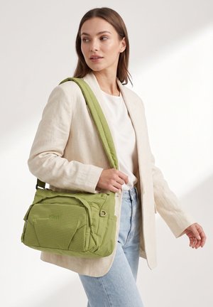 Femme portant un blazer beige et un jean bleu clair porte un sac à bandoulière vert clair avec plusieurs compartiments.