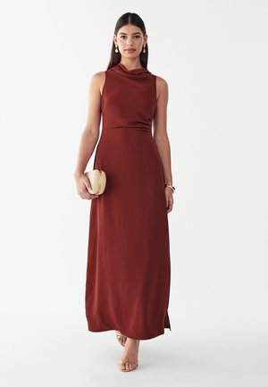 Frau mit langen dunklen Haaren trägt ein ärmelloses, rostfarbenes Maxikleid, beige Sandalen, Perlenohrringe und hält eine kleine beige Clutch.
