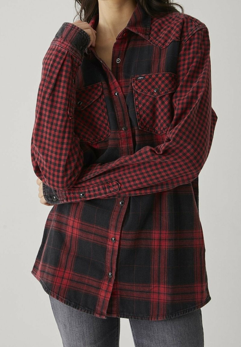 Personne portant une chemise à manches longues à carreaux rouge et noir avec des poches poitrine, debout les bras croisés, sur un fond neutre.