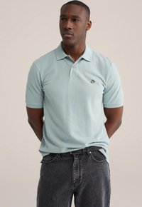 Lichtblauw katoenen poloshirt met een zwart logo op de linkerborst; voorzien van een kraag en twee knopen, gecombineerd met donkergrijze denim jeans.