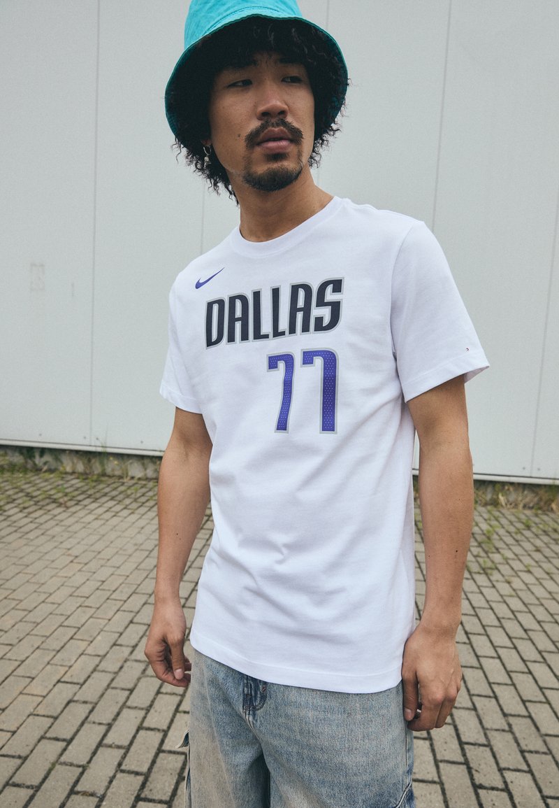 Hvid kortærmet t-shirt med "DALLAS" og nummer "77" i sort og lilla. Sammen med en lys blå bucket hat og denim shorts.