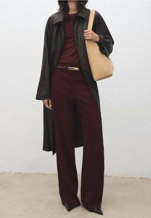 Czarny długi płaszcz na bordowym topie i szerokich bordowych spodniach. Nosi beżową torebkę na ramię. Czarne szpiczaste buty.