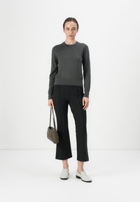 Grijze aangesloten trui, op maat gemaakte zwarte broek met een voornaad, witte instap schoenen en een kleine olijf-groene tas. Glad textuur over het geheel.