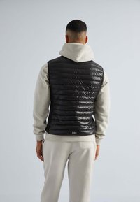 Gilet noir rembourré avec un matelassage horizontal, porté par-dessus un sweat à capuche gris clair. Doté d'un col haut et d'un logo en bas. Texture lisse et brillante.