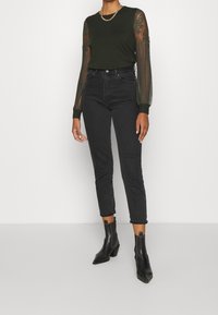 Donkergroene trui met doorzichtige puffmouwen, gecombineerd met hoge zwarte spijkerbroeken en zwarte puntige enkellaarsjes. Inclusief een zilveren ketting.