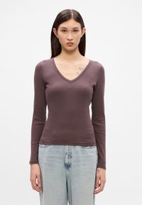 Top cu mâneci lungi, de culoare mauve, cu un decolteu în V și un accent mic cu logo pe piept, asortat cu blugi deschiși la culoare, ușor uzati.