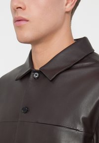 Sötétbarna bőrkabát, kiterjesztett gallérral, gombos záródással és sima textúrával, enyhe fényességgel.