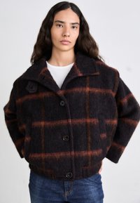 Giacca di lana a scacchi nera e arancione con un ampio colletto, quattro bottoni e una vestibilità rilassata, indossata sopra una camicia bianca e jeans di denim.