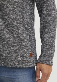 Leichter, strukturierter Pullover aus schwarz-weißer Mélange, mit einem kleinen braunen Lederetikett am Saum und schmalen Ärmeln.